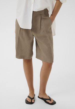 Persoon die losse bruine knielange shorts draagt, een wit overhemd met knopen en zwarte slippers, staand met één hand in de zak.