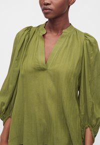 Femme portant un chemisier ample vert olive à col en V avec des manches longues bouffantes et froncées terminées par des poignets élastiques.