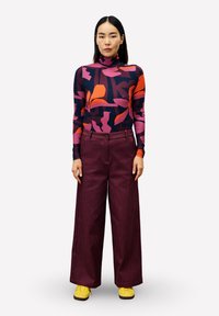 Femme debout portant un col roulé à motifs floraux à manches longues, un pantalon large bordeaux et des baskets jaunes sur un fond blanc.