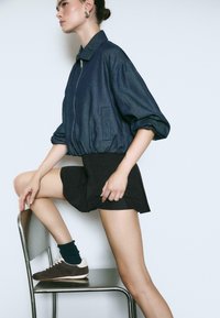 Denim-Bomberjacke mit elastischem Saum, Ballonärmeln und seitlichen Taschen, kombiniert mit schwarzen Shorts und braunen Turnschuhen. Das Model posiert mit einem Bein auf einem Stuhl.
