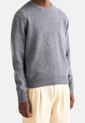 Hombre con suéter de lana gris claro y pantalones plisados beige, mostrado desde el cuello hasta las caderas sobre un fondo blanco.
