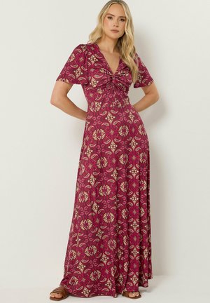 Kobieta o długich blond włosach, ubrana w czerwoną, wzorzystą suknię sięgającą ziemi, z krótkimi rękawami i marszczonym dekoltem w kształcie litery V, stojąca z rękami na biodrach.