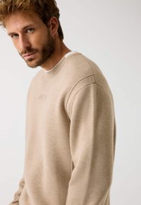 Maglione beige realizzato in tessuto morbido, con scollatura a girocollo, polsini a coste e un discreto dettaglio del logo in rilievo sulla parte anteriore.