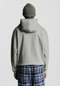 Persona che indossa un berretto in maglia olive, una felpa grigia chiara e pantaloni a quadri blu e neri, in piedi di fronte a uno sfondo grigio chiaro semplice.