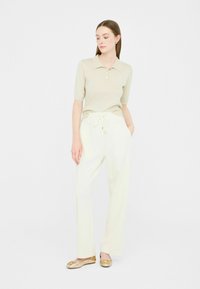 Polo shirt beige chiaro con maniche corte e tre bottoni, abbinato a pantaloni bianchi con cintura elastica e tasche laterali. Ballerine dorate.