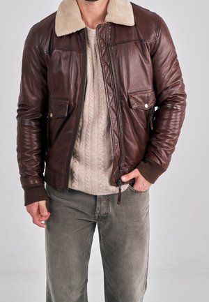 Uomo che indossa una giacca di pelle marrone con colletto in shearling sopra un maglione beige a trecce e pantaloni grigi, con la mano in tasca.