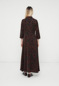 Femme aux longs cheveux blonds portant une robe maxi à fleurs foncées et des chaussures à lacets bordeaux brillantes, se tenant devant un fond blanc uni.