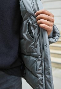 Jachetă căptușită gri cu un interior texturat și fermoar securizat. Dispune de un buzunar lateral și un material exterior durabil.