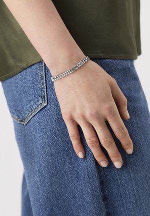 Main reposant sur la poche d'un jean bleu, portant un bracelet en argent ornés de strass en double rangée, avec une chemise vert olive visible au-dessus.