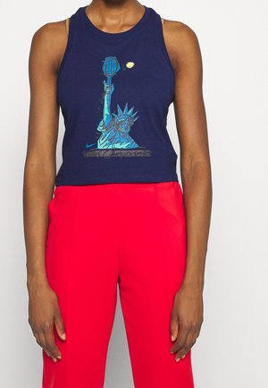 Person iført en marineblå ærmeløs croptop med grafik af Frihedsgudinden, der holder en tennisracket, kombineret med lyse røde bukser.