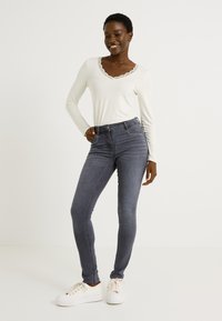 Camiseta blanca de manga larga con escote de encaje, combinada con jeans skinny gris oscuro y zapatillas blancas. Diseño sencillo con acentos mínimos.
