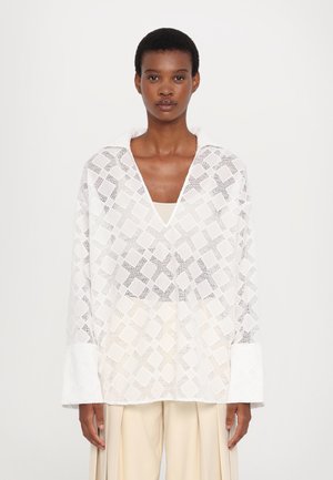 White, textured long-sleeve blouse with a loose fit, featuring a diamond pattern and a V-neckline, paired with beige pleated trousers.