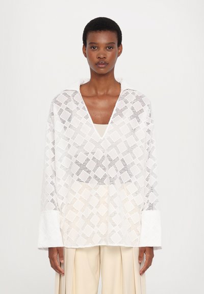 White, textured long-sleeve blouse with a loose fit, featuring a diamond pattern and a V-neckline, paired with beige pleated trousers.