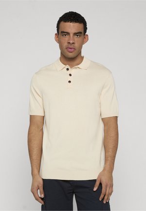 Młody mężczyzna z krótkimi kręconymi włosami, ubrany w beżową koszulkę polo z krótkim rękawem i ciemne spodnie, stojący na tle gładkiego jasnego tła.