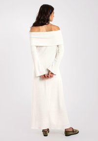 Ein schulterfreies, langärmliges weißes Kleid aus weichem, strukturiertem Stoff, mit einem taillierten Oberteil und ausladenden Ärmeln, kombiniert mit grünen Schuhen.