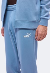 Personne portant un pantalon de survêtement Puma bleu clair et une veste assortie avec fermeture éclair, affichant le logo Puma sur la cuisse gauche et une main détendue sur le côté.