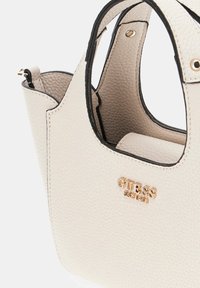 Beige színű, textúrázott kézitáska fekete díszítéssel, arany "GUESS" logóval. Kerek formájú, rövid fogantyúkkal, belső részletek láthatóak.