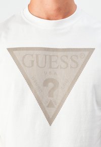 T-shirt blanc avec un logo Guess triangulaire marron comportant des lignes verticales et un point d'interrogation au centre, porté par une personne.