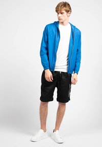 Blå letvægts hættetrøje med lynlås foran; parret med sorte opsmøgede shorts med grafiske detaljer på siden; hvide sneakers afslutter looket.