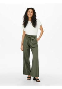 White short-sleeve top paired with green wide-leg pants featuring a tie waist. Model stands in black heeled sandals, smiling.