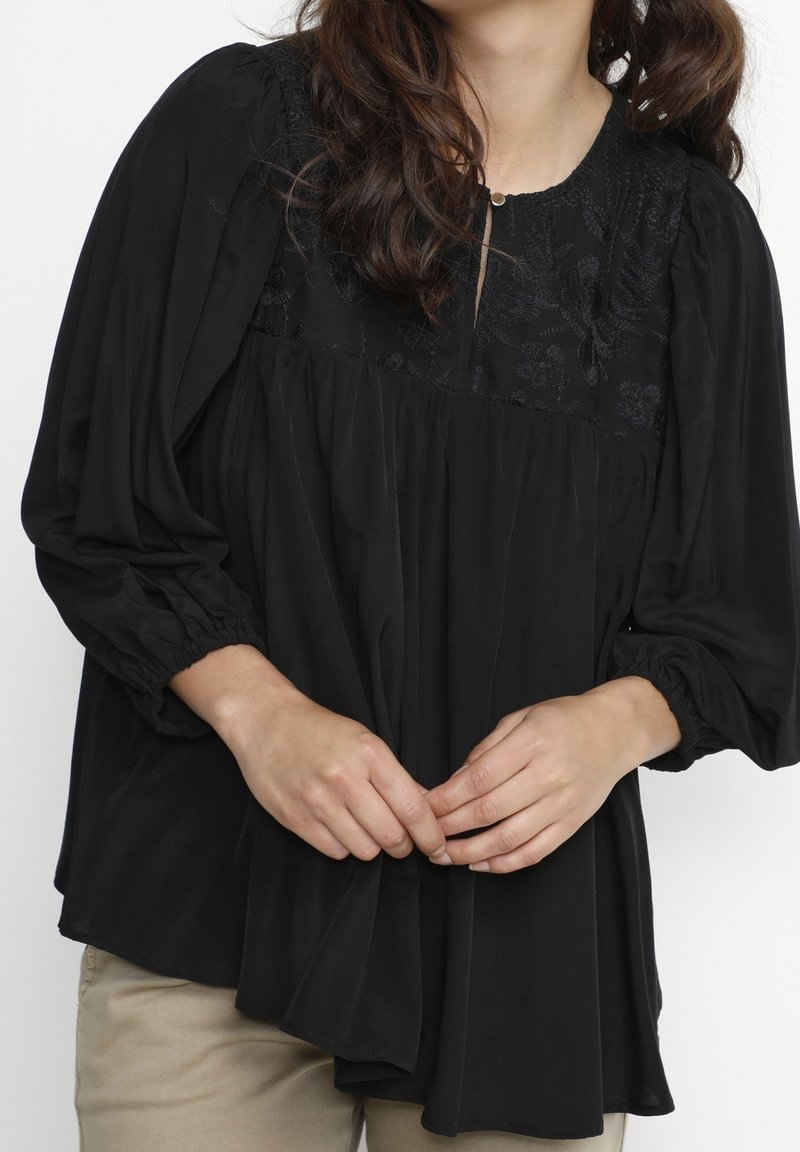 Svart blus med broderad byst, långa puffärmar med elastiska manschetter, samlad struktur och knäppning vid halsen.