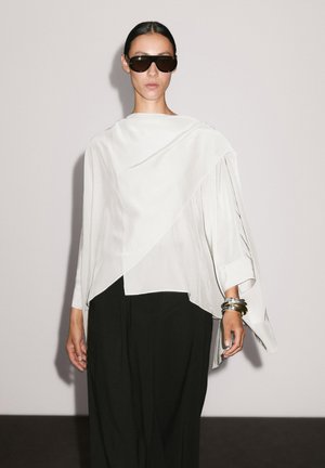 Übergroße weiße Bluse mit Drapierung und asymmetrischem Schnitt, kombiniert mit lockeren schwarzen Hosen. Das Model trägt dunkle Sonnenbrillen und silberne Armreifen.