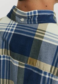 Camisa de cuadros para hombre con patrones de cuadros en azul, blanco y beige. Fabricada con una tela texturizada, cuenta con un cuello con botones y costuras visibles.