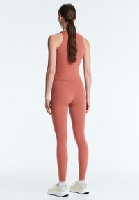 Femme aux longs cheveux bruns attachés en queue de cheval, portant un haut sans manches couleur rouille et des leggings, debout les mains détendues le long du corps, vue de dos.