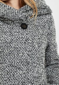 Donna che indossa un cappotto lavorato a maglia grigio e bianco con un colletto alto e un bottone scuro visibile vicino al collo.