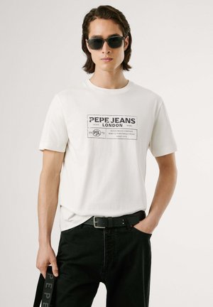 Witte t-shirt met korte mouwen en een zwart grafisch logo. Gecombineerd met zwarte jeans en een zwarte riem, model draagt donkere zonnebril.