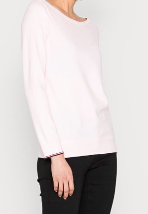 Personne portant un haut rose clair à manches longues et un pantalon noir, debout devant un fond blanc uni.