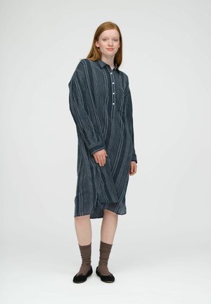 Jeune femme aux cheveux roux portant une robe bleue à motifs, manches longues, longueur genou, des chaussettes marron et des ballerines noires, debout sur un fond blanc uni.