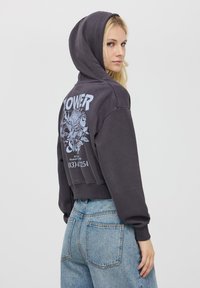 Przycięta czarna bluza z kapturem z niebieskim nadrukiem na plecach. Posiada kaptur i ściągacze rękawów, w połączeniu z jasnoniebieskimi dżinsami.