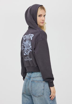 Felpa nera corta con stampa grafica blu sul retro. Presenta un cappuccio e polsini a coste, abbinata a jeans in denim azzurro chiaro.