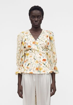 Γυναίκα που φοράει μία κρεμ υφασμάτινη μπλούζα με floral μοτίβο και 3/4 φουσκωτά μανίκια, καθώς και ανοιχτόχρωμο, φαρδύ παντελόνι, με φόντο απλό.