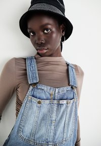 Šviesiai mėlyni džinsiniai kombinezonai su variniais priedais ir didelėmis kišenėmis, derinami su prigludusia smėlio spalvos ilgomis rankovėmis palaidine ir juoda kibiro skrybėle.