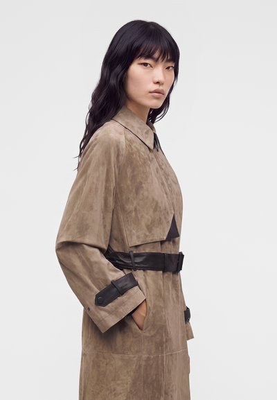 Jeune femme aux longs cheveux foncés portant un trench-coat beige en daim avec une ceinture en cuir noir et des détails aux poignets, debout devant un fond uni.