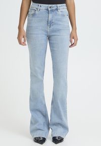 Vrouw draagt lichtblauwe jeans met hoge taille en wijde pijpen, en zwarte schoenen met puntige neus en zilveren gespen, tegen een witte achtergrond.