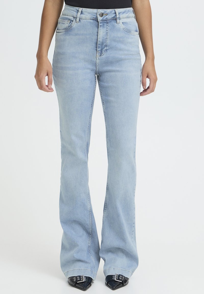 Vrouw draagt lichtblauwe jeans met hoge taille en wijde pijpen, en zwarte schoenen met puntige neus en zilveren gespen, tegen een witte achtergrond.