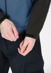 Langärmlige Jacke mit blauen Rumpf und schwarzen Ärmeln, mit Klettverschlüssen an den Bündchen und einer glatten Textur, getragen über dunklen Cargo-Hosen.