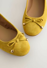 Ballerines en daim jaune avec bouts arrondis, nœuds décoratifs noués avec des embouts métalliques dorés, et semelles intérieures beige rembourrées sur un fond clair.