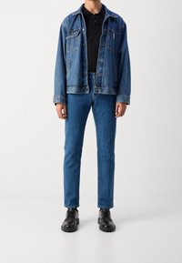 Chaqueta vaquera de color azul claro, con dos bolsillos en el pecho y cierre de botones, llevada sobre un polo negro y combinada con jeans ajustados azules y zapatos negros.