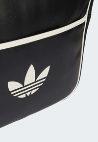 Juodo odinio krepšio, kuriame yra baltas Adidas logotipas su trimis lapų formos žiedlapiais ir dviem horizontaliomis juostelėmis, kraštai aptraukti balta siūle.