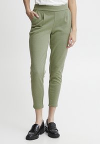 Groene, aansluitende broek met een gladde textuur, hoge tailleband en voorzakken. Draagt een paar zwarte, glanzende loafers.