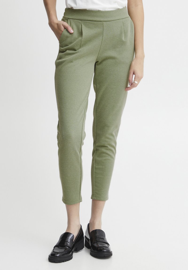 Groene, aansluitende broek met een gladde textuur, hoge tailleband en voorzakken. Draagt een paar zwarte, glanzende loafers.