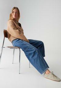 Pull beige épais, pantalon ample en jean bleu et baskets gris clair. Assis sur une chaise chromée devant un fond uni.