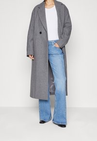 Abrigo de lana gris con un único botón negro, que presenta un corte relajado y grandes solapas, combinado con unos pantalones vaqueros de campana de color azul claro y zapatos negros.