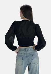 Schwarze transparente Bluse mit langen Ärmeln und gerafftem Bund, kombiniert mit hellblauen, hochtaillierten Jeans mit Gesäßtaschen.