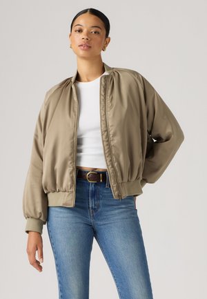 Femme portant une veste bomber marron clair sur un t-shirt court blanc avec un jean bleu et une ceinture marron, debout sur un fond uni.