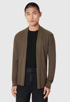 Ung mand med kort hår iført en brun åben cardigan over en sort skjorte og mørke jeans mod en ensfarvet baggrund.
